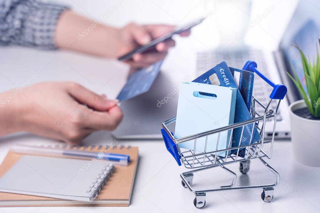 Online paying shopping, electronic payment with credit card, smart phone concept, laptop over white table background with shop cart trolley, close up.