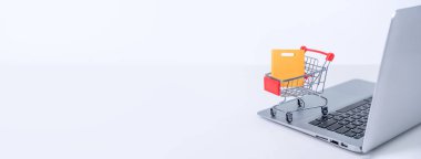 İnternetten alışveriş. Beyaz masa arkaplanındaki dizüstü bilgisayarın üzerinde renkli kağıt torbalarla mini market arabası, ev konseptiyle satın alma, kapat.