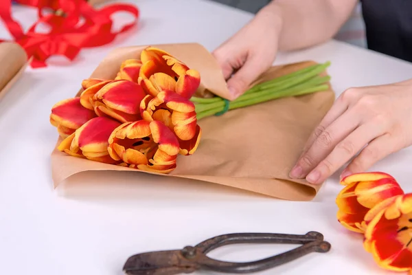 Young female florist is wrapping a red color tulip flower bouquet for Mother\'s Day gift with tie ribbon bow over a white table background, close up, lifestyle.