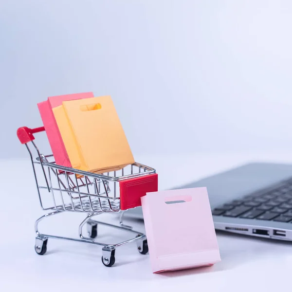 Online shopping. Mini shop cart trolley with colorful paper bags over a laptop computer on white table background, buying at home concept, close up