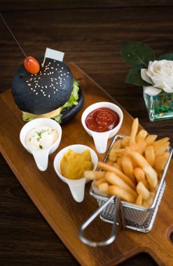 Koyu ahşap masa arka plan üzerinde siyah burger gemide. Siyah hamburger eti patty, peynir, domates, mayonez, patates kızartması bir kağıt bardak ile. Modern hızlı yiyecek öğle yemeği
