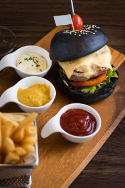 Koyu ahşap masa arka plan üzerinde siyah burger gemide. Siyah hamburger eti patty, peynir, domates, mayonez, patates kızartması bir kağıt bardak ile. Modern hızlı yiyecek öğle yemeği