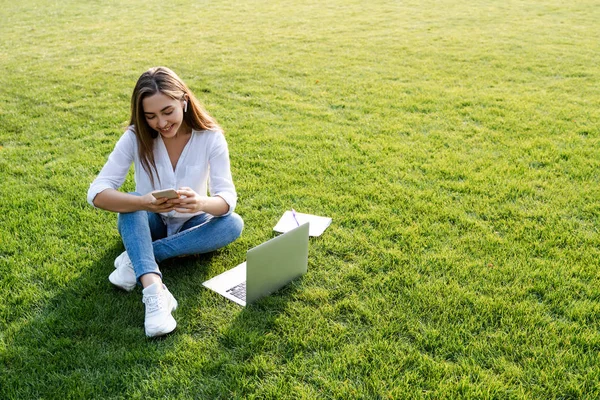通过搜索手机和使用笔记本电脑 让积极的女学生或年轻学生坐在公园外的绿草上 自由职业或学习的概念 有复制空间 生活方式 — 图库照片