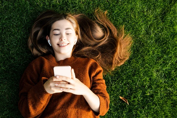 微笑着快乐的少女看着她的智能手机 打电话给朋友聊天 在网上交流 利用现代数字在线和网络技术 放松地躺在绿草上 — 图库照片