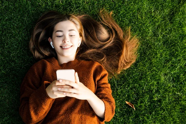 Gülümseyen mutlu genç kız akıllı cep telefonuna bakıyor ve arkadaşlarıyla çevrimiçi iletişim kurmak için konuşuyor, modern dijital online ve internet teknolojisini kullanıyor, yeşil çimlerin üzerinde uzanarak rahatlıyor