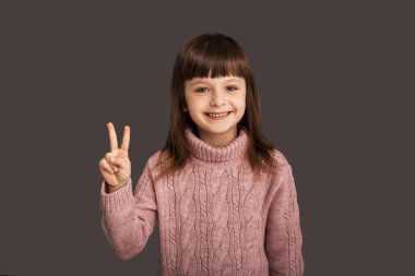 Herkese barış ve sevgi, gri arka plandaki tatlı esmer kız iki parmağını kaldırmış, nazik ve mutlu çocuğu gösteriyor.