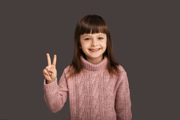 Herkese barış ve sevgi, gri arka plandaki tatlı esmer kız iki parmağını kaldırmış, nazik ve mutlu çocuğu gösteriyor.