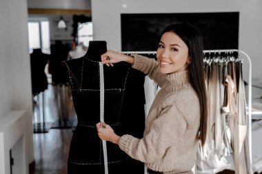 Önlüklü esmer terzi atölyedeki siyah kukla üzerinde güzel kumaş ölçüyor. Kameraya gülümseyen, Asyalı profesyonel terzi ustası. 