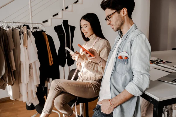 In the studio of tailoring and sewing clothes, a pair of young tailors, before starting work, look at social networks on phones and communicate online on the Internet