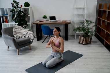 Genç bir kadın namaste jesti ile yoga yapıyor, yoga egzersizi yapıyor, asana pozu veriyor, antrenman yapıyor, spor kıyafeti giyiyor, gri pantolon giyiyor, spor sutyen giyiyor, ev dekorasyonu yapıyor.