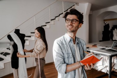 Genç yaratıcı insanlar yeni bir kıyafet koleksiyonu yapmakla meşguller, ön planda yakışıklı bir adam. Tasarım, dikiş ve kesim stüdyosu, tasarımcı giysileri, imalat, el işi ürünleri.
