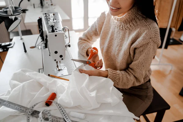 Genç dahi terzilik stüdyosundaki dikiş makinesinin yanındaki masada beyaz bir bezi sürüyor ve kesiyor. Gülümsüyor ve yeni bir tasarımcı kıyafeti oluşturma sürecinden zevk alıyor.