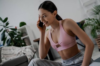 Sabah egzersizi, bir kadın evde yerde egzersiz yapmadan önce oturur, telefonda mutlu bir şekilde konuşur ve gülümser, spor bir bluz ve eşofmanla. Sağlıklı yaşam tarzı, spor çalışmaları.