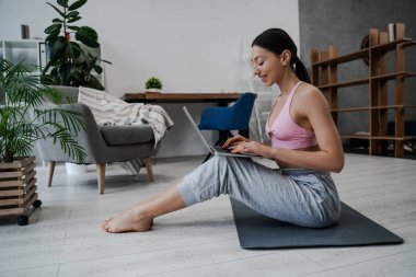 Genç atletik atletik kız sabah egzersizlerinden önce spor minderinin üzerinde yerde oturur, bazı kas grupları üzerinde egzersiz yapar, dizüstü bilgisayarda esneme ve yoga asanalarını arar. 