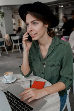 Dikey görüntü. Telefondan konuşan, şapkalı ve son moda yeşil tişörtlü bir kadın. Serbest çalışan, girişimci, sindirme alanı, kamusal alan, modern insan yaşam tarzı, iş çevrimiçi konsepti.