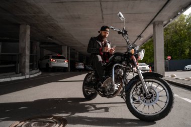 Motosikletli, motorsikletli, siyah sakallı şık hippi sakallı motosikletli adam, cep telefonu kullanan acımasız adam ve internetteki 5G internet bağlantıları. İnsan yaşam tarzı ve hobisi