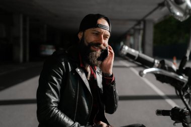 5g internet bağlantısı kullanıcısı. Motosikletli gezgin, vahşi sakallı sokak yarışçısı motosikletinde oturuyor ve telefonla konuşuyor. İnsanların yaşam tarzı ve hobileri. Teknoloji konsepti.