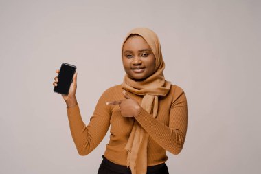 Telefonu gösteriyor ve beyaz stüdyo arka planında geleneksel tesettürlü Afrikalı Müslüman kadını gösteriyor. İnsanların yaşam tarzı. Dünya kültürleri. Reklam resmi