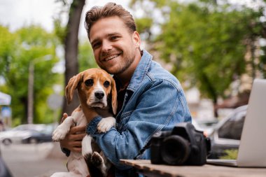 Hayvanlar ve köpekli genç ve mutlu adamı sokak kafesinde geziyor, internette dizüstü bilgisayarda çalışıyor, mesafesinde özgürce çalışıyor. Mutlu arkadaşlar, hayvan bakımı ve sevgi.