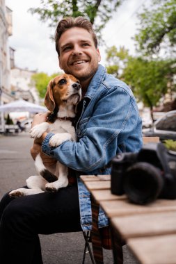 Dikey görüntü mutlu serbest fotoğrafçı köpeğiyle kafede oturuyor ve rahatlamak ve eğlenmek için zaman buluyor. Havalı gülümseyen hippi, modern adam, teknoloji kullanıcısı.