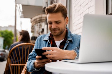 Genç hippi blog yazarı cep telefonunu tut ve dışarıda iletişim için dizüstü bilgisayarı kullan. Caddedeki kafede oturup, gülümseyip, mutlu olmak. Çevrimiçi arama ve internet teknolojisi kavramı