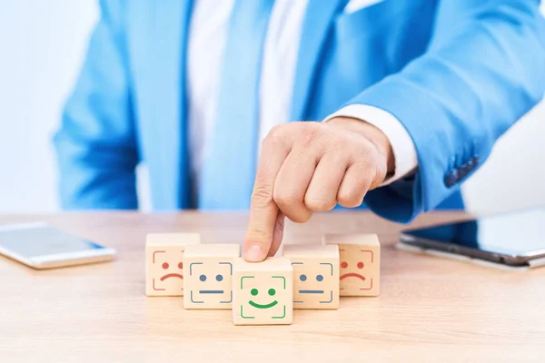 The concept of satisfaction assessment. Six cubes lie in a pyramid on the businessman\'s table, with icons of various emotions on the cubes. The man advances the central cube with an emotion of joy.