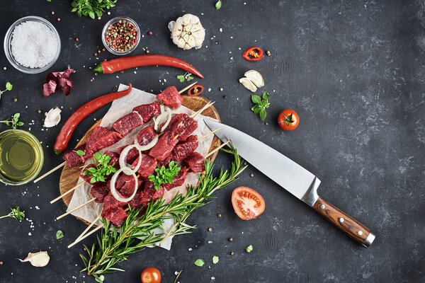 Five bamboo skewers with fresh beef meat lie on parchment and a cutting board on a dark background. Seasonings, spices, vegetables and herbs are on the table. Top view, free space for text.