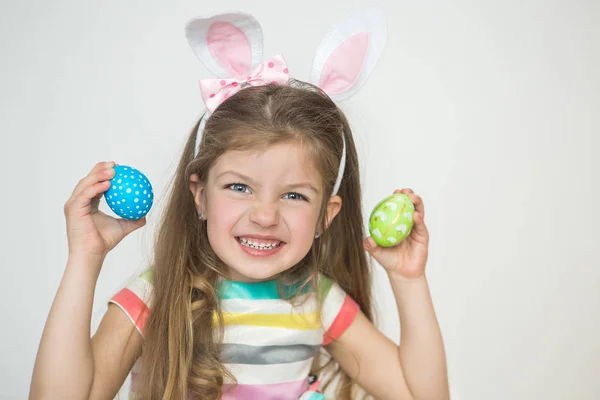 Tavşan kulağı takan, elinde boyalı Paskalya yumurtaları tutan ve beyaz arka planda gülümseyen sevimli küçük bir kız. Paskalya Günü.