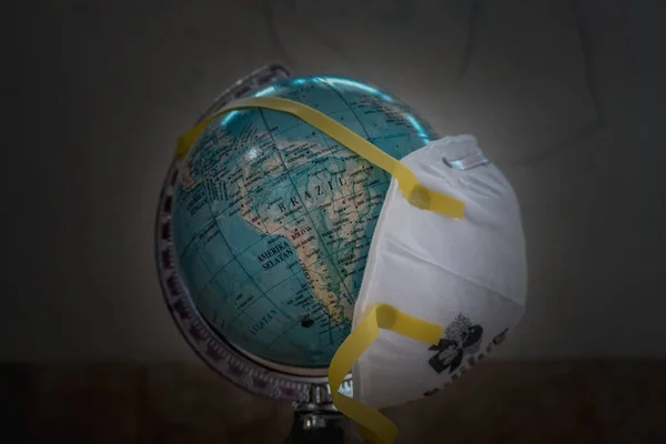 a mock earth globe covered with an N95 mask, signifying the world being attacked by the covid-19 pandemic. Dark background helps focus on globe objects and n95 masks