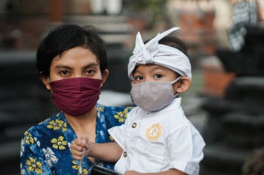 Balili anne ve çocuk kordemik ya da covid-19 sırasında geleneksel Bali kıyafetleri giyiyorlar. İkisi de kendilerini virüs saldırılarından korumak için maske kullanıyor..