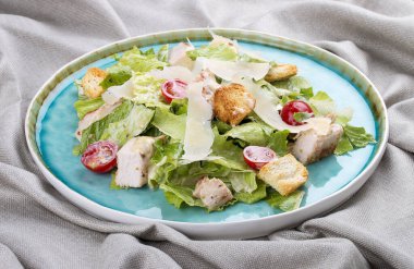 Tavuk, parmesan ve vişneli domatesli Sezar salatası. Tekstil arka planında mavi bir tabakta