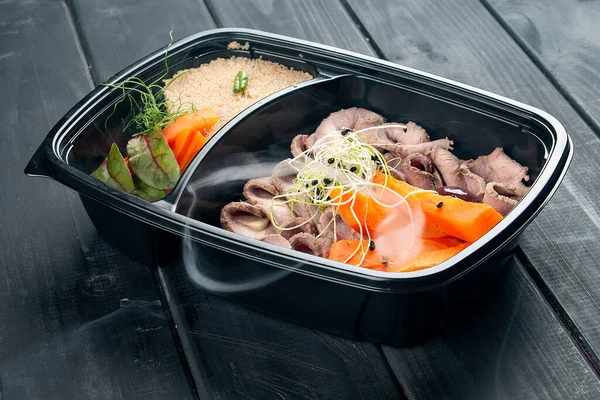 Ham with couscous and vegetables in containers on a black wooden background. Takeaway. Diet and healthy food.