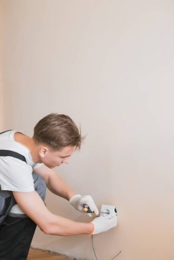 Genç gülümseyen elektrik teknisyeni soket mekanizmasını beyaz bir duvardaki kablolara bağlıyor. Tornavida kullanıyor, yeni bir dairede tamirat yapıyor.