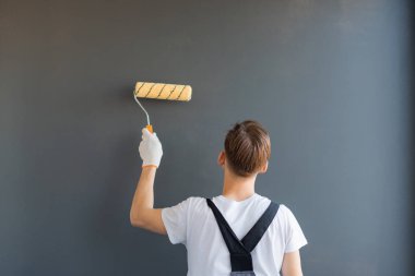 Tulum giymiş yakışıklı genç bir işçi sırtı dönük duruyor ve duvarları gri bir arka planla boyuyor.