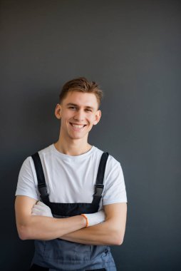 Gri tulumlu, beyaz tişörtlü, gri arka planda kolları çapraz duran genç, güler yüzlü, yakışıklı, profesyonel bir işçinin portresi. Dikey resim, metin için yer, kopyalama alanı
