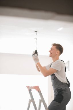 Tulumlu, elinde tornavidayla evin içinde lamba tamir eden genç bir elektrikçi. Ev onarımı konsepti. Metin için boşluk.