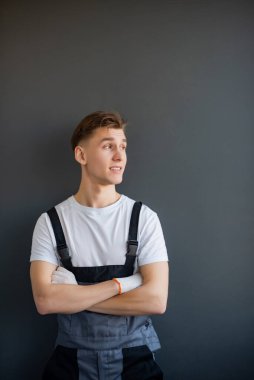 Gri tulumlu, beyaz tişörtlü, gri arka planda kolları çapraz duran genç, güler yüzlü, yakışıklı, profesyonel bir işçinin portresi. Dikey resim, metin için yer, kopyalama alanı