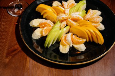 Meyve salatası karışımı, meyve tatlısı dilimlenmiş muz, mandalina, portakal, elma. Siyah tabakta meyveler. Lezzetli ve sağlıklı bir tatlı.