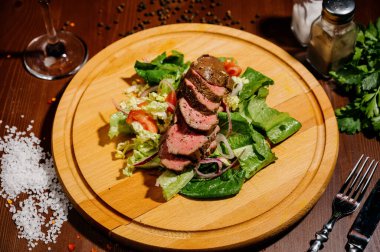 Ahşap bir tahtanın üzerinde salatalı bir jambon tabağı. Masanın arkasında koyu bir arka plan var.