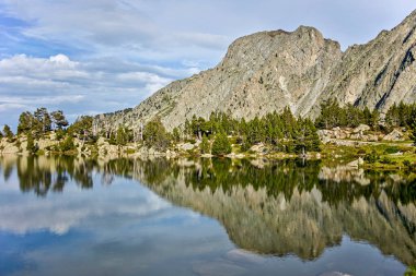 Ağaçlardan ve dağlardan oluşan bir gölde yansıması olan ve beyaz bulutlu mavi bir gökyüzü olan ideal dağ manzarası.