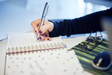 Kırmızı tırnaklı ve siyah kazaklı bir kadın eli resmi. Metal halkalı bir deftere bir çift gözlük ve bilgisayar faresinin yanında yazıyor..