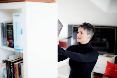 Stüdyosunda kısa saçlı, siyah giyinmiş bir kadın yatak odasında kanepesi ve masasında küresi olan beyaz bir kitaplıkta kitaplara bakıyor.