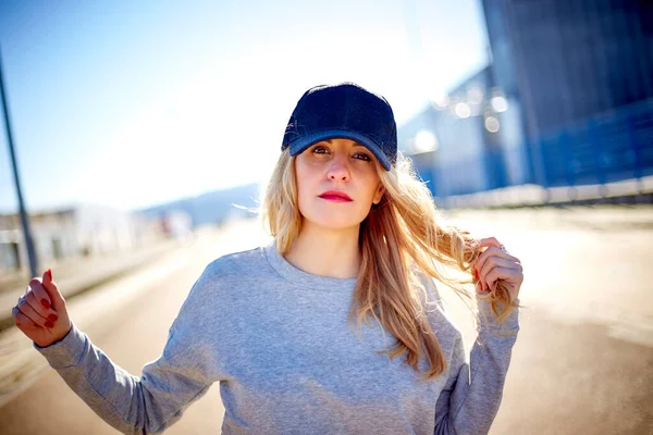Girl with a cap and long blonde hair, wearing a grey sweatshirt, walking down the street in an empty urban area on a very sunny day and playing with her hair