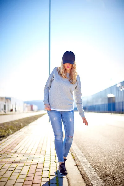 Girl with cap and long blonde hair, wearing jeans, sneakers and sweatshirt, walking down the street in an empty urban area on a very sunny day
