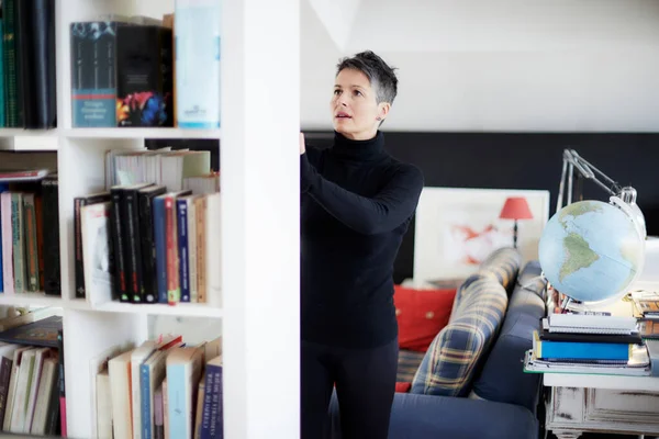 Woman dressed in black with short hair in her home studio looking at books in a white bookcase with a couch in the bedroom and a globe on the desk