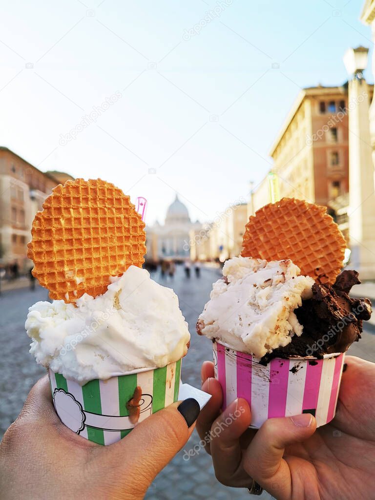 An image of two paper cups with an ice cream of various sorts in the hands of a girl and a man, who are on the street on the background of the city.