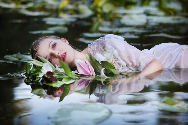 ユリの花束を持つ白いドレスで水に横たわっている美しいセクシーな女性 — ストック写真