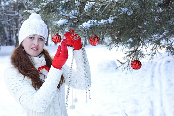 Όμορφο κορίτσι σε ένα λευκό πουλόβερ και καπέλο διακοσμεί ένα κλαδί πεύκου με κόκκινες μπάλες Χριστουγέννων — Φωτογραφία Αρχείου