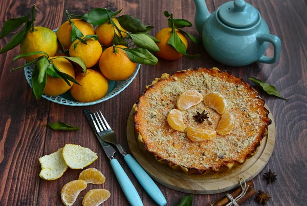 Вид на вкусный цитрусовый пирог, корзина с сочными мандаринами, чайник, вилка и нож на коричневом деревянном столе — стоковое фото