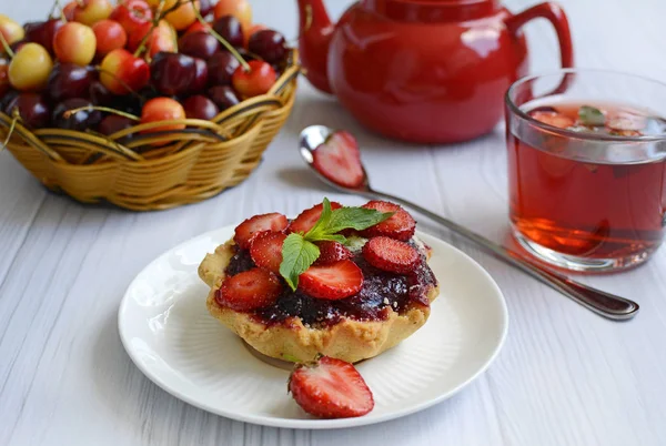 Appetitliche Cupcake mit Erdbeeren, rote Teekanne, Tasse und Korb mit reifen Kirschen auf einem hellen Holztisch — Stockfoto